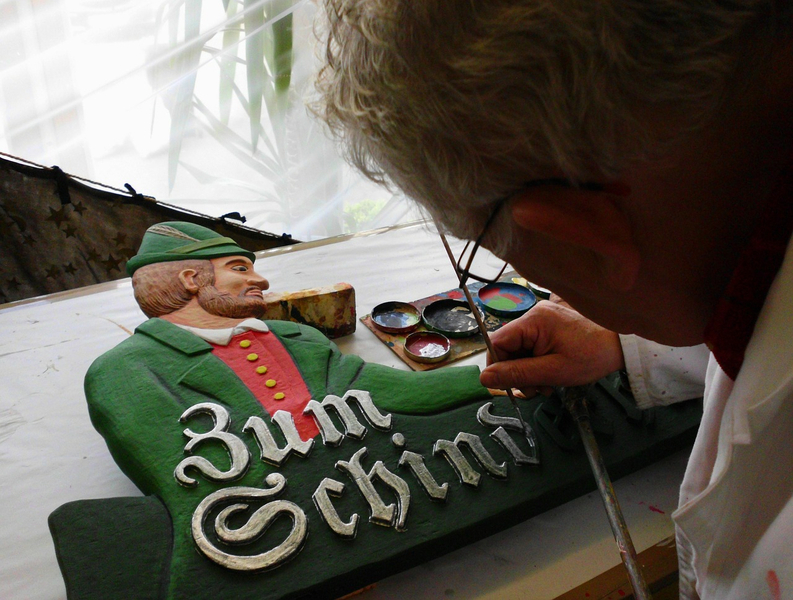 Mensch bearbeitet ein historisches Holzschild eines Mannes in grüner Jacke mit Aufschrift: Zum Schind