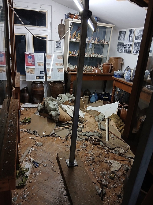 Blick in einen durch den Tornado schwer beschädigten Ausstellungsraum des Museums im Kannenofen in Höhr-Grenzhausen. Eine Metallstütze sichert die beschädigte Decke; Trümmer und Dämmmaterial liegen zwischen Keramikgefäßen und Vitrinen auf dem Boden.